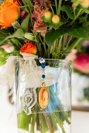 Glass vase with colorful flowers, lace ribbon, and a gold cameo pendant with blue beads.