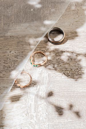 Three wedding rings on a rustic wooden surface with a sheer veil, featuring gold bands with emerald accents and a silver ring.