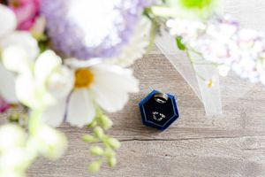 Blue velvet hexagon ring box with wedding rings on wooden surface surrounded by white and purple flowers.