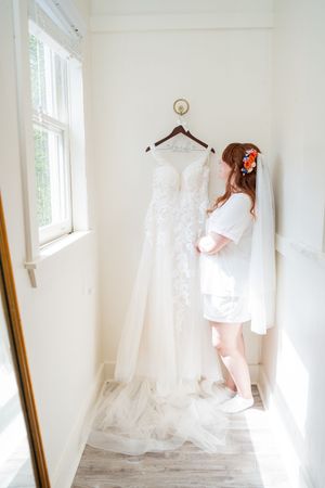 Bride admiring her white lace wedding gown hanging by a sunlit window before her ceremony.