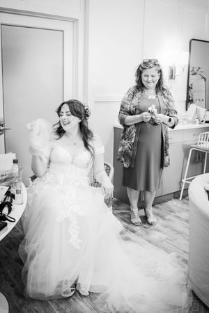 Black and white photo of a bride in a lace gown laughing with a woman in a dress during wedding preparations.