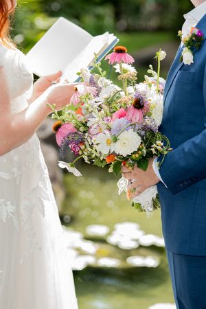 Bride reading vows holding colorful wildflower bouquet with pink coneflowers and white dahlias, groom in navy suit beside pond.