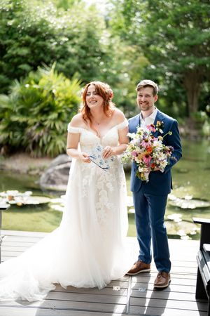 Smiling bride in off-shoulder lace gown and groom in navy suit stand on dock holding colorful bouquet at garden wedding.