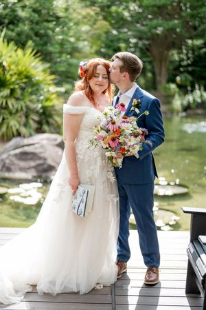 Bride and groom portrait on a garden dock, she holds a wildflower bouquet as he kisses her cheek.