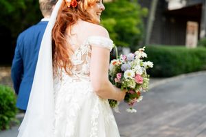 Red-haired bride in lace wedding dress holding colorful bouquet, viewed from behind.