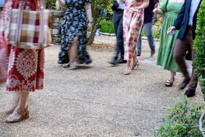 Wedding guests in colorful dresses and heels mingle on gravel garden path during reception.