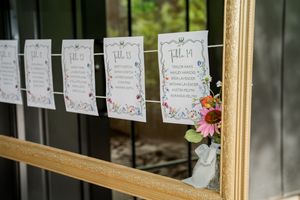 Wedding seating chart cards with floral borders hanging on twine in a gold frame, decorated with fresh flowers.