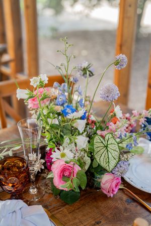 Colorful wildflower wedding centerpiece with pink roses, blue delphiniums, and lush greenery on a rustic wooden table.