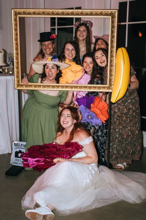 Bride and bridesmaids posing playfully inside a gold picture frame at a wedding reception photo booth.
