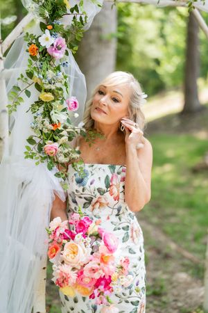 Elegant older woman in floral dress holding colorful bouquet beside flower-adorned arch outdoors.