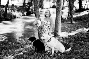 Black and white photo of a smiling bride holding flowers, posing outdoors with two dogs by trees.