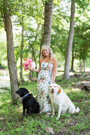 Smiling blonde bride in floral gown holds pink bouquet beside black and yellow Labrador dogs in lush green forest.