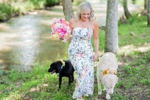 Blonde woman in floral jumpsuit holding pink bouquet walks two flower-crowned dogs by a creek outdoors.