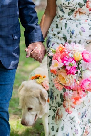 Couple holding hands at wedding with golden retriever dog and colorful pink floral bouquet, bride in floral dress.