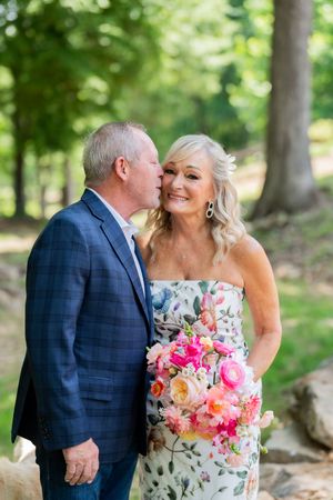 A man in a navy blazer kisses a smiling blonde woman in a floral dress holding a pink bouquet outdoors.