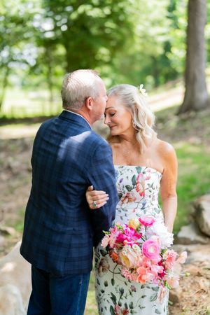 Older couple sharing a romantic moment outdoors, bride in floral dress holding pink bouquet, groom in navy plaid suit.