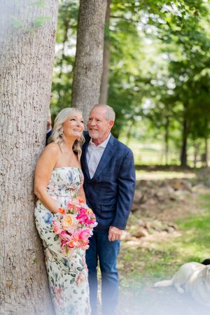 A couple in formal attire poses by a tree in a lush garden, woman in floral dress holding a colorful bouquet.