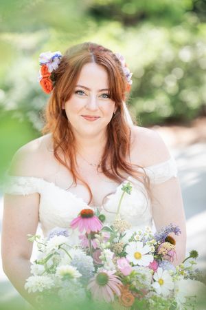Radiant bride with red hair and floral crown holds wildflower bouquet in white off-shoulder gown outdoors.