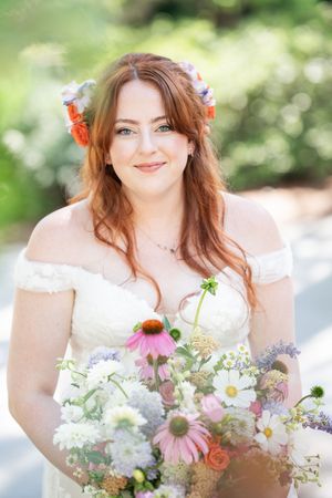 Smiling redhead bride holding a wildflower bouquet with echinacea and daisies, wearing a floral crown outdoors.