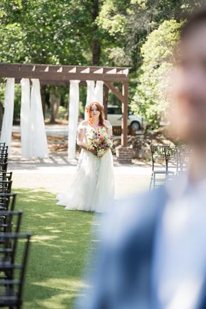 Red-haired bride in white gown walking down outdoor aisle holding colorful bouquet.