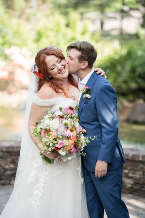 Smiling bride holding colorful bouquet receives kiss on cheek from groom in blue suit outdoors.