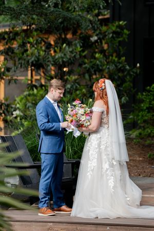 Bride and groom exchange vows outdoors, she holds colorful bouquet in lace gown, he wears blue suit, lush greenery background.