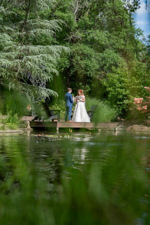 Bride and groom stand together on a wooden dock surrounded by lush greenery reflected in a pond.