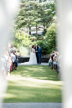 Outdoor wedding ceremony on green lawn aisle with seated guests and lush garden backdrop.