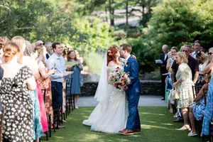 Bride and groom share a kiss at outdoor wedding ceremony while guests line the aisle cheering.