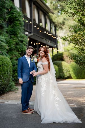 Bride in lace gown and groom in blue suit pose together outside elegant venue with string lights.