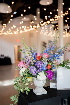 Colorful wedding floral arrangement in white urn vase with string lights bokeh background.