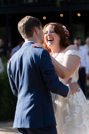 Joyful bride in lace gown laughs while sharing first dance with groom in navy suit.