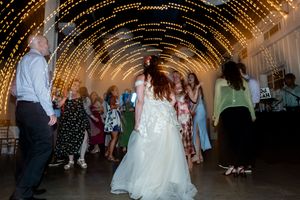 Bride dances with guests under arch of twinkling lights at lively wedding reception.