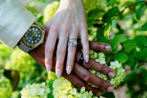 Two hands stacked on white hydrangeas, one wearing a diamond ring and one a gold watch, symbolizing love and commitment.