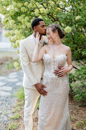 Interracial couple shares a tender kiss in garden, groom in beige suit, bride in beaded strapless gown.