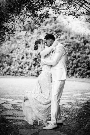 Black and white photo of bride and groom embracing tenderly under garden arbor on their wedding day.