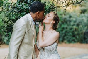 Interracial couple sharing a romantic kiss in a garden, groom in beige suit, bride in strapless lace wedding gown.