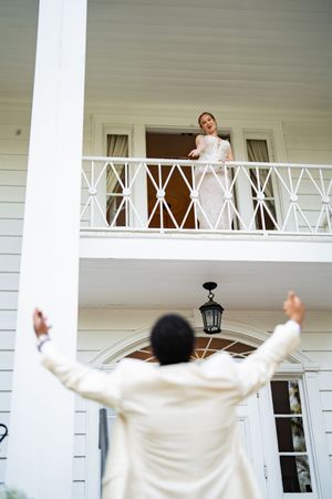 Groom in white suit raises arms toward bride on balcony of elegant white colonial mansion.