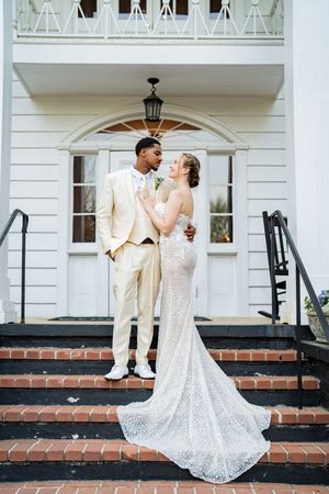 Interracial couple poses on brick steps outside white church, groom in cream suit, bride in lace mermaid wedding gown.