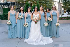Bridesmaids in dusty blue dresses holding white bouquets pose with bride in lace gown at outdoor wedding venue.