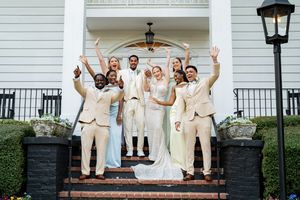 Wedding party in cream suits and pastel dresses celebrating on brick steps outside a white mansion.