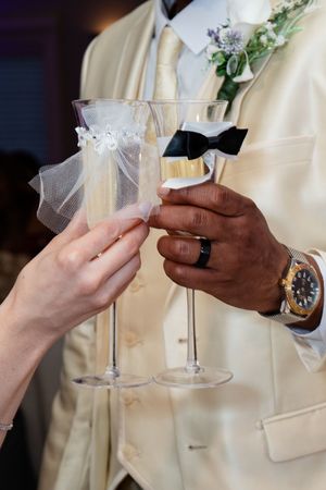 Interracial couple clinking decorated champagne flutes at wedding reception, groom in cream tuxedo with bow tie.