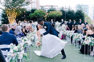 Wedding guests seated at outdoor garden reception with white chairs and floral decorations at sunset.