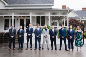 A group of groomsmen lined up in front of a white farmhouse wearing coordinated navy and gray suits.