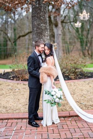 Bride in white gown with fur stole and veil holds bouquet while groom in tuxedo embraces her on brick path.