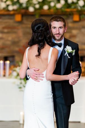 Bride and groom share their first dance, groom in black tuxedo with boutonniere, bride in elegant backless white gown.
