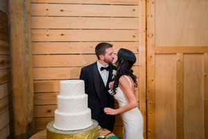 Bride and groom share a kiss beside a three-tier white wedding cake in a rustic barn venue.
