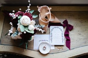Wedding details flatlay with pink shoes, floral bouquet, and invitation cards on wooden surface.