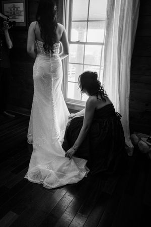 Black and white photo of a wedding dress being adjusted near a bright window.
