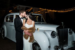 Bride and groom share a romantic kiss beside a classic white Rolls-Royce at night, with string lights glowing above.